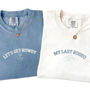 May include: Two t-shirts, one light blue and one white, with gold necklaces. The light blue shirt says "LET'S GET ROWDY" with an anchor graphic. The white shirt says "MY LAST RODEO" with a cowboy hat graphic.