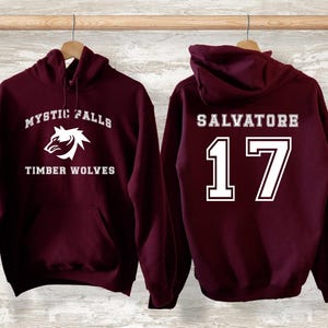 May include: Two maroon hoodies hanging on a wooden rod. One displays "MYSTIC FALLS TIMBER WOLVES" with a wolf graphic. The other has "SALVATORE 17" in white lettering. Both have a hood and a front pocket.