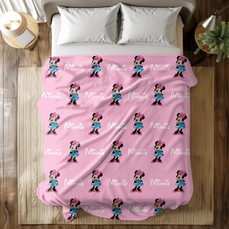 Puede incluir: Manta rosa de Minnie Mouse con un patr&oacute;n repetido de Minnie Mouse con vestido y lazo azules, y la palabra "Minnie" en blanco. La manta est&aacute; sobre una cama con almohadas blancas.