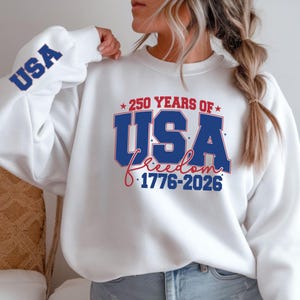 May include: White sweatshirt with the text "250 YEARS OF USA Freedom 1776-2026" in blue and red. The word "USA" is also printed in blue on the left sleeve.