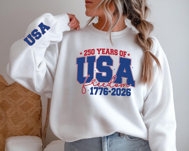 May include: White sweatshirt with the text "250 YEARS OF USA Freedom 1776-2026" in blue and red. The word "USA" is also printed in blue on the left sleeve.