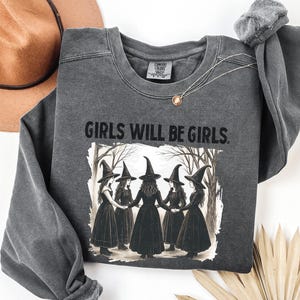 May include: Dark gray sweatshirt with the text "GIRLS WILL BE GIRLS" above a graphic of five witches holding hands in a forest. The witches are wearing black dresses and pointed hats. A brown hat is in the background.
