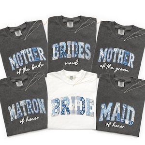 May include: A collection of gray and white t-shirts with text in a collegiate style. The shirts say "Mother of the Bride", "Bridesmaid", "Mother of the Groom", "Matron of Honor", "Bride", and "Maid of Honor". The text is in blue and white floral patterns.