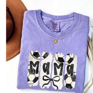 Puede incluir: Una camiseta lila Comfort Colors con la palabra "Mama" en negro y gr&aacute;ficos de balones de f&uacute;tbol. Se ve un collar dorado y un sombrero marr&oacute;n. La camiseta tiene cuello redondo.