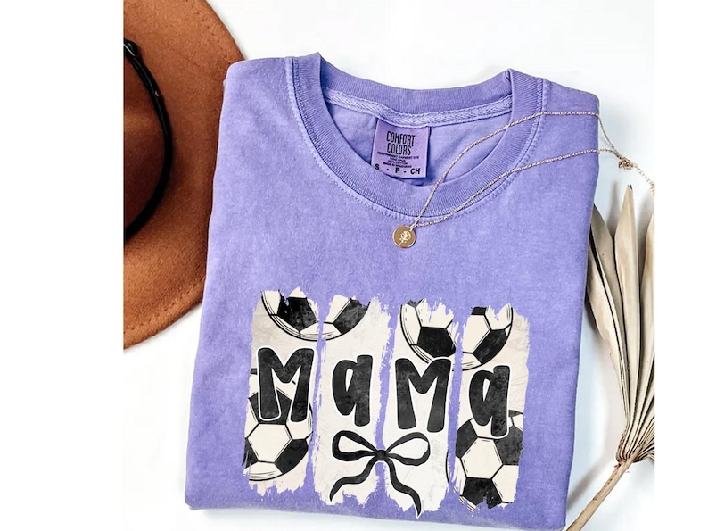 Puede incluir: Una camiseta lila Comfort Colors con la palabra "Mama" en negro y gr&aacute;ficos de balones de f&uacute;tbol. Se ve un collar dorado y un sombrero marr&oacute;n. La camiseta tiene cuello redondo.
