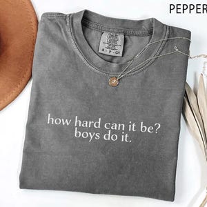 May include: A grey Comfort Colors t-shirt with the slogan "how hard can it be? boys do it." A silver necklace with a pendant rests on the shirt. A brown hat and dried plant are in the background.