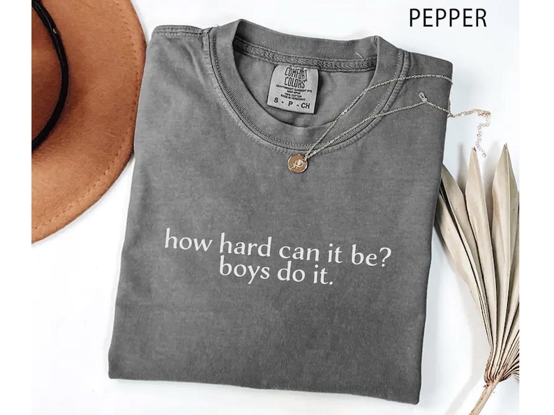 May include: A grey Comfort Colors t-shirt with the slogan "how hard can it be? boys do it." A silver necklace with a pendant rests on the shirt. A brown hat and dried plant are in the background.