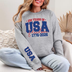 May include: A gray sweatshirt with the text "250 YEARS OF USA freedom 1776-2026" in red and blue. The sleeve has the word "USA" in blue. The sweatshirt is worn by a person.