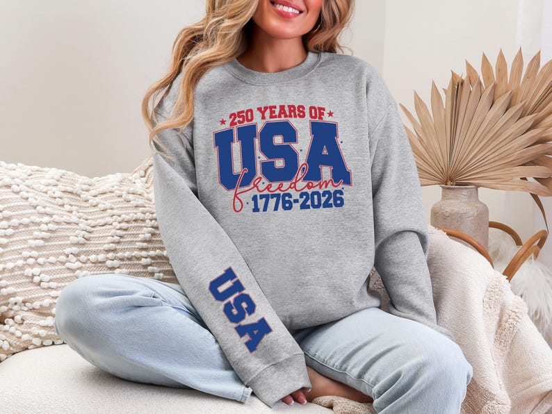 May include: A gray sweatshirt with the text "250 YEARS OF USA freedom 1776-2026" in red and blue. The sleeve has the word "USA" in blue. The sweatshirt is worn by a person.