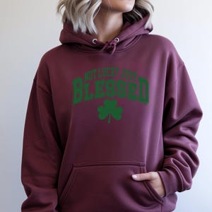 May include: Burgundy hoodie with a front pocket and drawstring hood. The hoodie features the text "NOT LUCKY JUST BLESSED" in green, arched across the top, with a green shamrock below. A casual, comfortable garment.