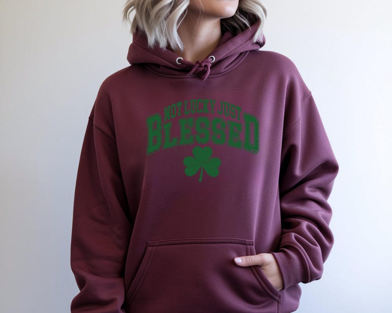 May include: Burgundy hoodie with a front pocket and drawstring hood. The hoodie features the text "NOT LUCKY JUST BLESSED" in green, arched across the top, with a green shamrock below. A casual, comfortable garment.