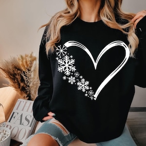 May include: A black long-sleeve sweatshirt featuring a large white graphic of a heart outline on the front. The left side of the heart is composed of various sizes of white snowflake designs, while the right side is a simple white line.