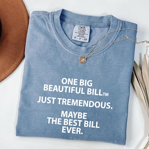 May include: A blue t-shirt with the text "ONE BIG BEAUTIFUL BILL, JUST TREMENDOUS, MAYBE THE BEST BILL EVER." A gold necklace with a pendant is on the shirt. The shirt is folded on a white surface.