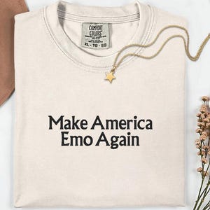 May include: Cream-colored t-shirt with the phrase "Make America Emo Again" in black lettering. A gold star pendant necklace rests on the shirt. The shirt has a Comfort Colors tag.