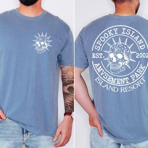 K&ouml;nnte beinhalten: Hellblaues Kurzarm-T-Shirt mit wei&szlig;em Totenkopf-Motiv auf der Vorder- und R&uuml;ckseite. Auf der R&uuml;ckseite des Shirts steht der Text "SPOOKY ISLAND AMUSEMENT PARK ISLAND RESORT EST. 2002" um ein Totenkopf-Design.