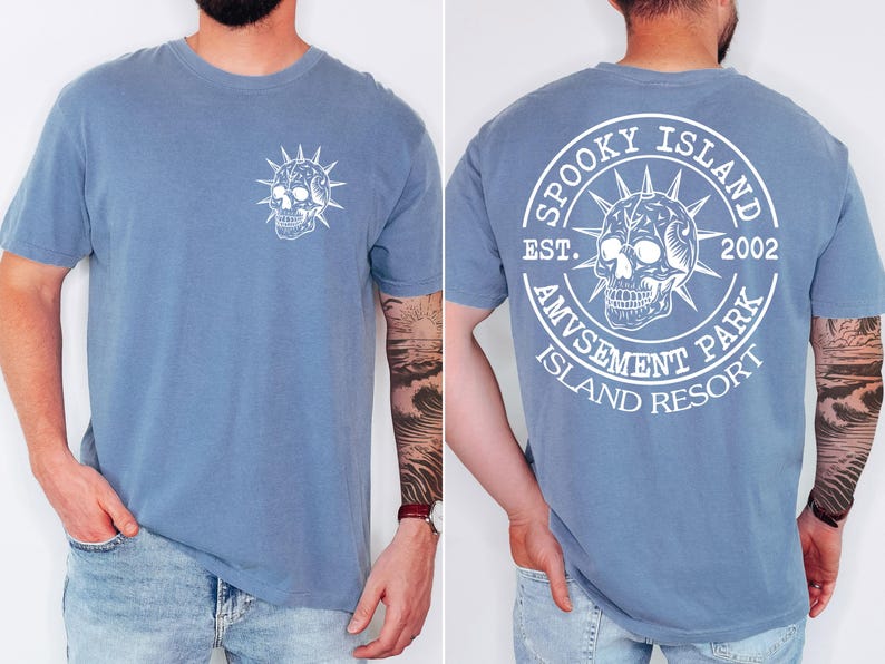 K&ouml;nnte beinhalten: Hellblaues Kurzarm-T-Shirt mit wei&szlig;em Totenkopf-Motiv auf der Vorder- und R&uuml;ckseite. Auf der R&uuml;ckseite des Shirts steht der Text "SPOOKY ISLAND AMUSEMENT PARK ISLAND RESORT EST. 2002" um ein Totenkopf-Design.