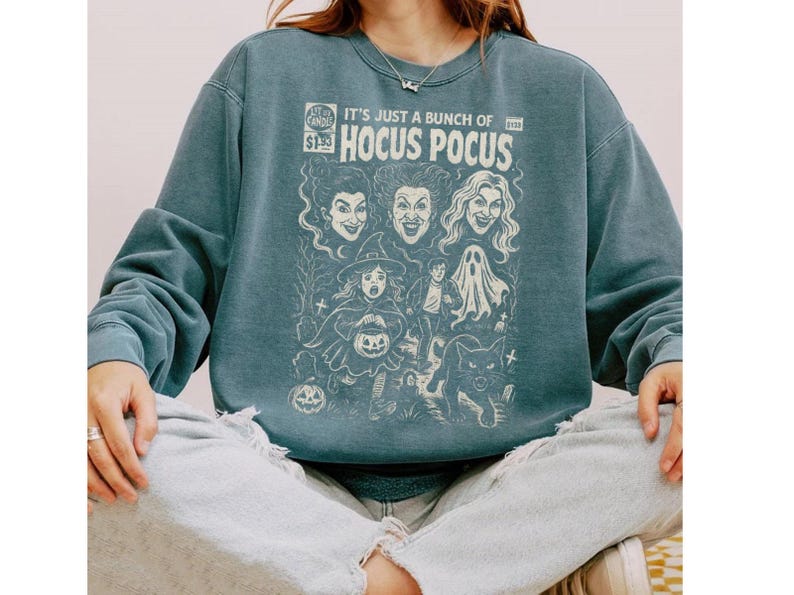 Może przedstawiać: Łupkowoniebieska bluza z okrągłym dekoltem i kremowym nadrukiem graficznym stylizowanym na okładkę komiksu vintage. Napis głosi IT'S JUST A BUNCH OF HOCUS POCUS, a całość uzupełniają ilustracje trzech kobiet, dziecka w kostiumie, ducha oraz kota.