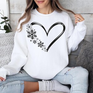 May include: A white long-sleeve sweatshirt featuring a black graphic print on the chest. The design consists of a large heart outline where the left side is composed of various sizes of black snowflakes, creating a winter-themed heart shape.