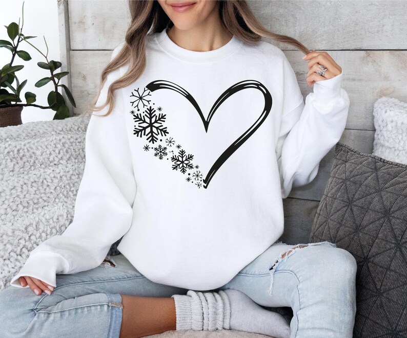 May include: A white long-sleeve sweatshirt featuring a black graphic print on the chest. The design consists of a large heart outline where the left side is composed of various sizes of black snowflakes, creating a winter-themed heart shape.