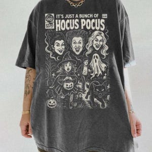 Może przedstawiać: Ciemnoszary t-shirt z kr&oacute;tkim rękawem z białym nadrukiem graficznym inspirowanym okładką komiksu. Wz&oacute;r zawiera napis "IT'S JUST A BUNCH OF HOCUS POCUS" oraz ilustracje trzech kobiet, dziecka w stroju czarownicy, ducha i kota.