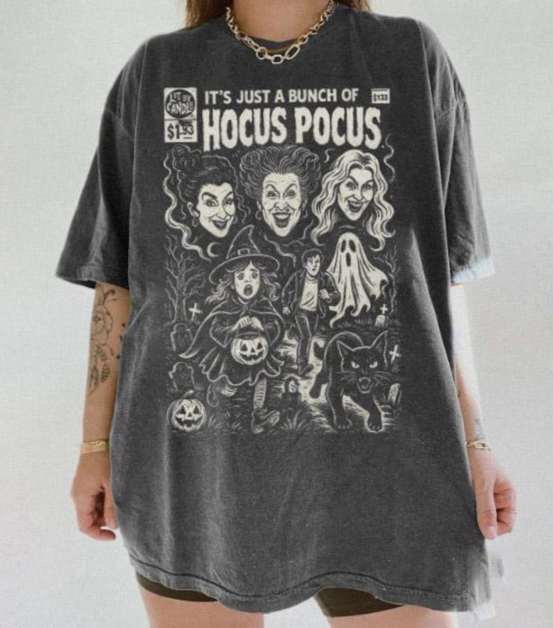 Może przedstawiać: Ciemnoszary t-shirt z kr&oacute;tkim rękawem z białym nadrukiem graficznym inspirowanym okładką komiksu. Wz&oacute;r zawiera napis "IT'S JUST A BUNCH OF HOCUS POCUS" oraz ilustracje trzech kobiet, dziecka w stroju czarownicy, ducha i kota.