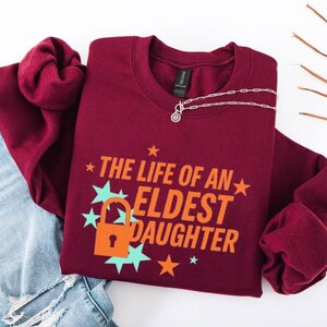 May include: A maroon crewneck sweatshirt with the text "THE LIFE OF AN ELDEST DAUGHTER" in orange and white. The design includes a lock and stars in teal and orange. A silver necklace with a pendant is on the sweatshirt.
