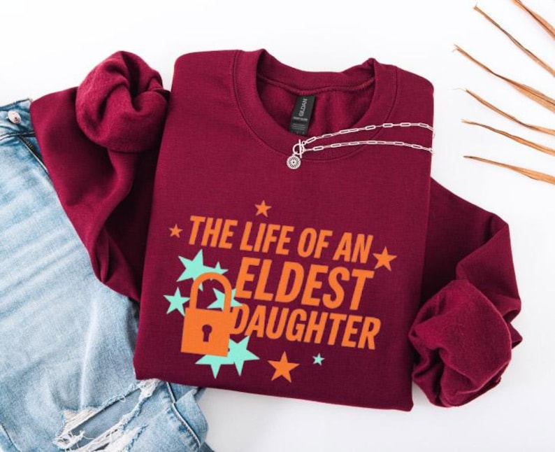May include: A maroon crewneck sweatshirt with the text "THE LIFE OF AN ELDEST DAUGHTER" in orange and white. The design includes a lock and stars in teal and orange. A silver necklace with a pendant is on the sweatshirt.