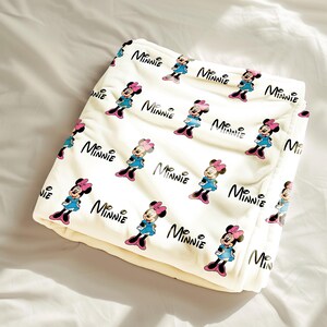 Puede incluir: Manta crema con un patr&oacute;n repetido de Minnie Mouse con vestido azul y lazo rosa, y la palabra "Minnie" en escritura negra. La manta est&aacute; doblada y parece suave.