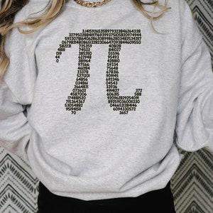 May include: A grey sweatshirt with black text printed on the front. The text includes a series of numbers, including 3.14159265358979323846264338, 3279502884197169399375105820974944, 592307816406286208998628034825342117, 067982148086513282306647093844609550, 408128, 841027, 55596, 95493, 428810, 59334, 75648, &pi;, 582231, 725359, 48111, 74502, 019, 3852110, 44, 622948, 0, 381964, 97566, 461284, 23378, 678316, 5271201, 909145, 64856, 034861, 692346, 04543, 2664821, 072602, 33936, 4914127, 4587006, 606315, 174881520, 715364367, 53054882, 951941511, 70, 372, 588, 920962829254091, 892590360011330, 0466521384146, 6094330572, 3657.