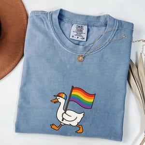 May include: A light blue Comfort Colors t-shirt featuring a cartoon goose wearing a rainbow cap and carrying a rainbow flag. The shirt has a crew neck and short sleeves. A gold necklace with a pendant is draped across the shirt.