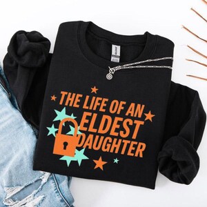 May include: Black sweatshirt with the text "THE LIFE OF AN ELDEST DAUGHTER" in orange, with a lock graphic. The design includes teal starbursts and orange stars. A silver necklace with a pendant is on the shirt.