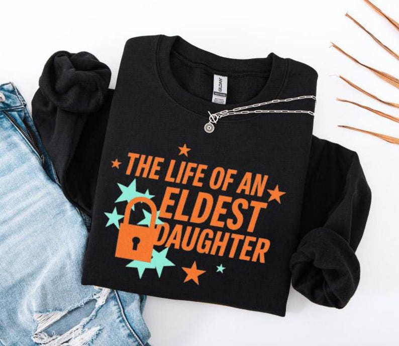 May include: Black sweatshirt with the text "THE LIFE OF AN ELDEST DAUGHTER" in orange, with a lock graphic. The design includes teal starbursts and orange stars. A silver necklace with a pendant is on the shirt.