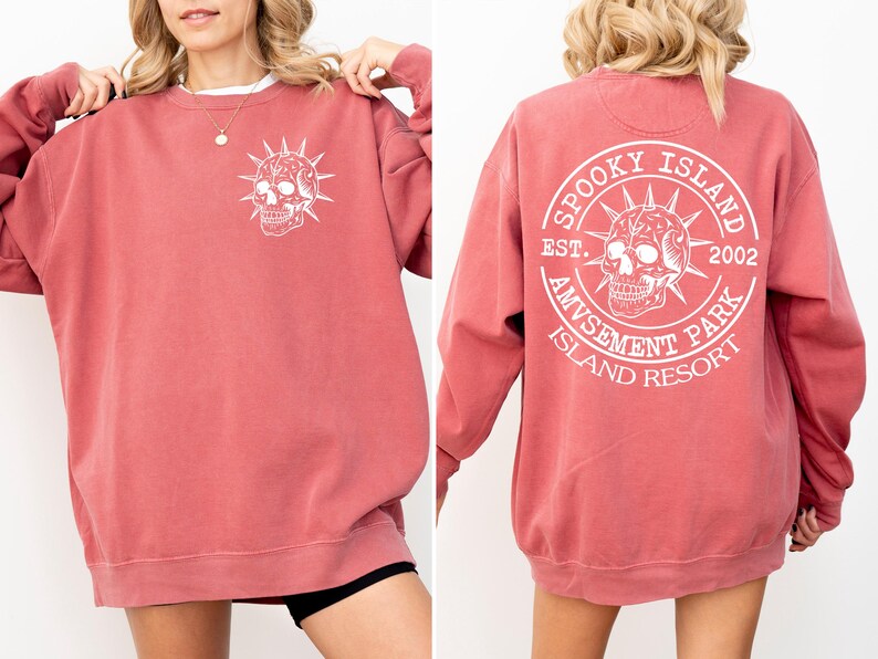 K&ouml;nnte beinhalten: Ein altrosa Sweatshirt mit einem wei&szlig;en Totenkopf-Motiv auf der Vorder- und R&uuml;ckseite. Die Vorderseite zeigt ein Totenkopf-Design, w&auml;hrend die R&uuml;ckseite ein kreisf&ouml;rmiges Design mit dem Text "Spooky Island Amusement Park Island Resort" und dem Jahr "2002" zeigt.