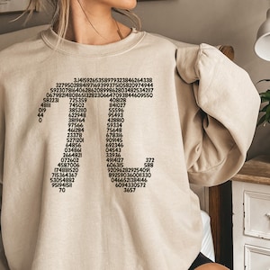 May include: A beige sweatshirt featuring a black graphic print of a cross formed from numbers. The numbers are arranged vertically, starting with 3.14159265358979323846264338 at the top and ending with 3657 at the bottom.