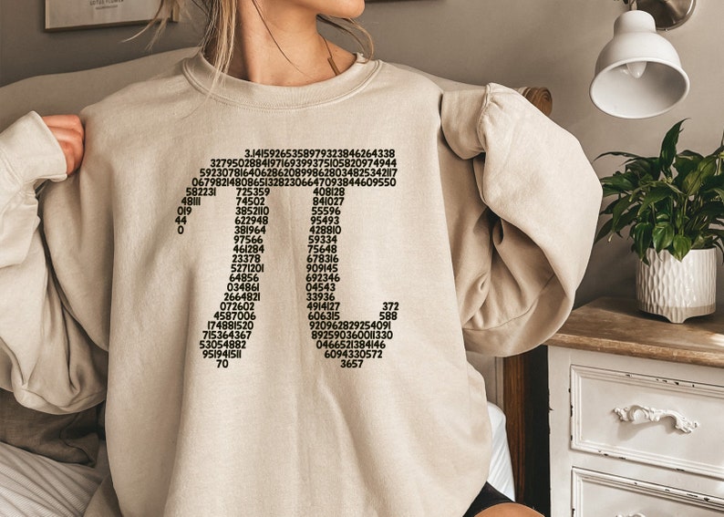 May include: A beige sweatshirt featuring a black graphic print of a cross formed from numbers. The numbers are arranged vertically, starting with 3.14159265358979323846264338 at the top and ending with 3657 at the bottom.