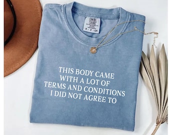 Può includere: Una t-shirt blu a girocollo con una finitura slavata. La parte anteriore presenta un testo bianco che recita: THIS BODY CAME WITH A LOT OF TERMS AND CONDITIONS I DID NOT AGREE TO. La maglietta ha un'etichetta del marchio Comfort Colors sul collo.