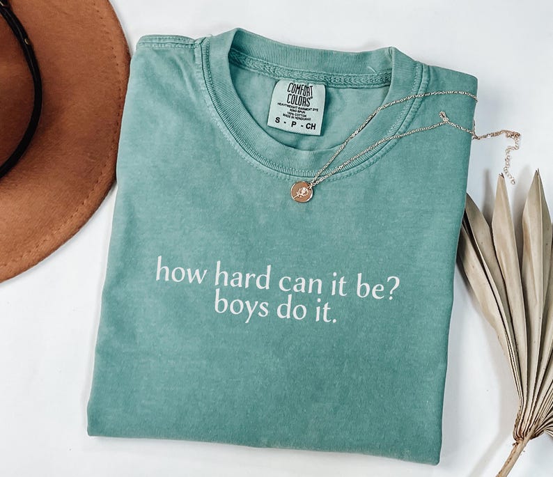 May include: A teal t-shirt with the slogan "how hard can it be? boys do it." A gold necklace with a circular pendant is on the shirt. The shirt is folded, and a brown hat is partially visible.