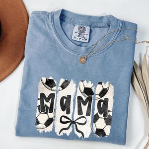 Puede incluir: Una camiseta azul Comfort Colors con la palabra "Mama" en fuente blanca desgastada, adornada con balones de f&uacute;tbol y un lazo. Un collar dorado con un colgante est&aacute; sobre la camiseta.