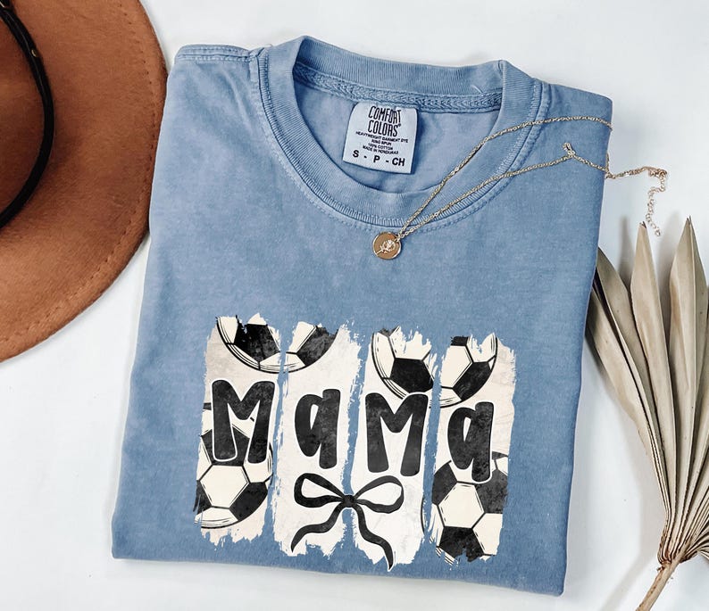 Puede incluir: Una camiseta azul Comfort Colors con la palabra "Mama" en fuente blanca desgastada, adornada con balones de f&uacute;tbol y un lazo. Un collar dorado con un colgante est&aacute; sobre la camiseta.