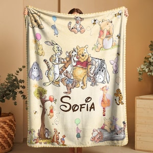 May include: A cream-colored blanket featuring illustrations of Winnie the Pooh and friends. The blanket has a yellow and white striped border and the name "Sofia" in brown script. Characters include Pooh, Tigger, Piglet, Eeyore, and Rabbit.