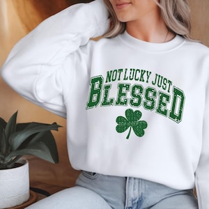 May include: White sweatshirt with the text "NOT LUCKY JUST BLESSED" in green, arched over a green shamrock. The sweatshirt is a crew neck style.