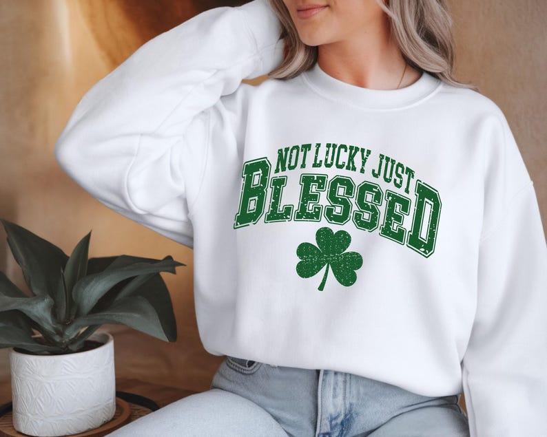 May include: White sweatshirt with the text "NOT LUCKY JUST BLESSED" in green, arched over a green shamrock. The sweatshirt is a crew neck style.