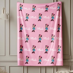 Puede incluir: Manta rosa con un patr&oacute;n repetido de Minnie Mouse con un vestido azul y un lazo rosa. La palabra "Minnie" est&aacute; impresa en blanco entre cada personaje. La manta est&aacute; colgada en una pared.