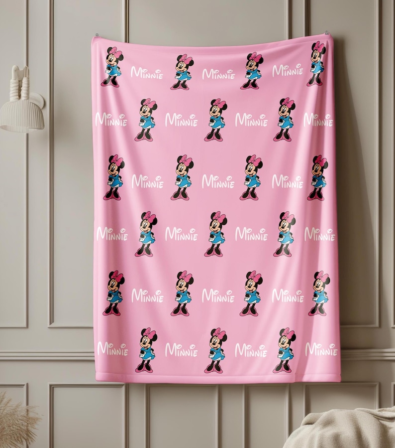 Puede incluir: Manta rosa con un patr&oacute;n repetido de Minnie Mouse con un vestido azul y un lazo rosa. La palabra "Minnie" est&aacute; impresa en blanco entre cada personaje. La manta est&aacute; colgada en una pared.
