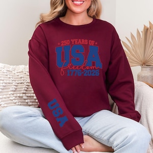 May include: Burgundy sweatshirt with the text "250 YEARS OF USA Freedom 1776-2026" in blue and red. The sleeve features the word "USA" in blue. The sweatshirt is a crew neck style.
