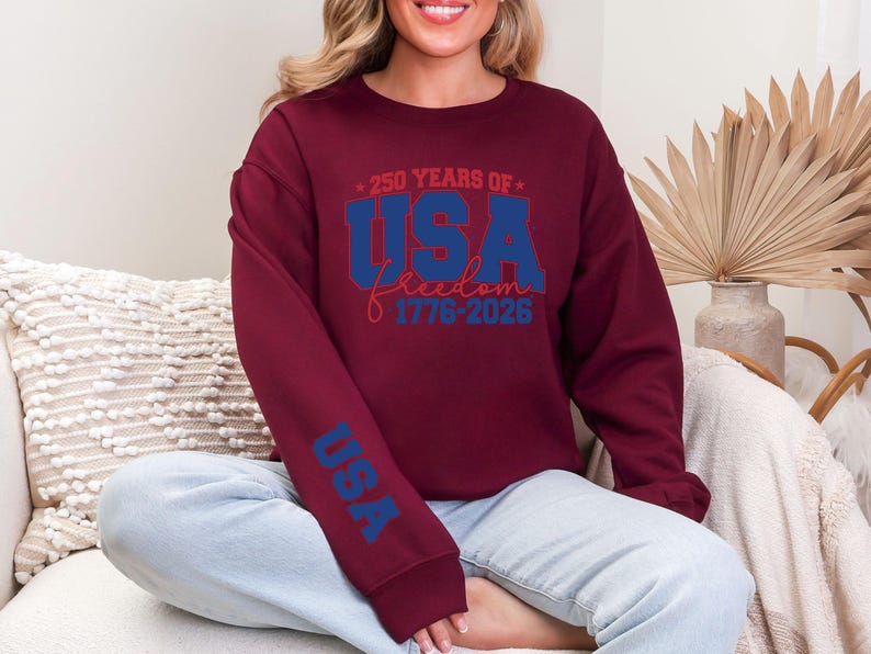 May include: Burgundy sweatshirt with the text "250 YEARS OF USA Freedom 1776-2026" in blue and red. The sleeve features the word "USA" in blue. The sweatshirt is a crew neck style.