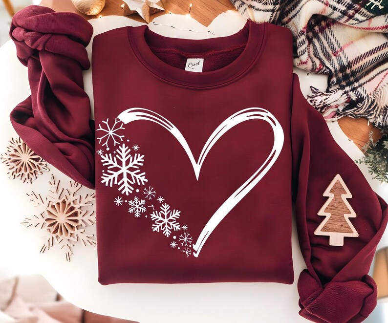 May include: A maroon long-sleeve sweatshirt featuring a large white graphic of a heart outline on the front. The left side of the heart is composed of various sizes of white snowflakes. The interior neck label displays the text "Cust".