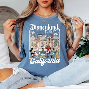 May include: A blue t-shirt featuring a Disneyland graphic with Mickey Mouse, Donald Duck, and other Disney characters. The graphic includes the text "Disneyland Est. 1955 California" in a vintage style. The shirt is a solid blue color.