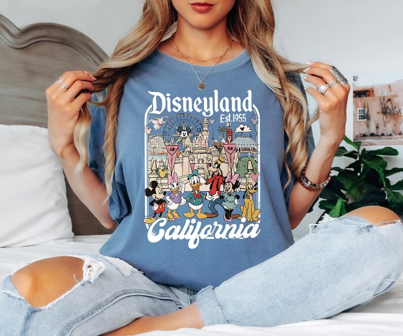 May include: A blue t-shirt featuring a Disneyland graphic with Mickey Mouse, Donald Duck, and other Disney characters. The graphic includes the text "Disneyland Est. 1955 California" in a vintage style. The shirt is a solid blue color.