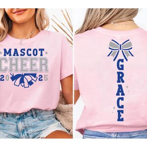 May include: Pink cheerleading t-shirt with the text "Mascot Cheer 2025" and a blue and white pom-pom. The back of the shirt has a silver bow and the text "GRACE" in blue.
